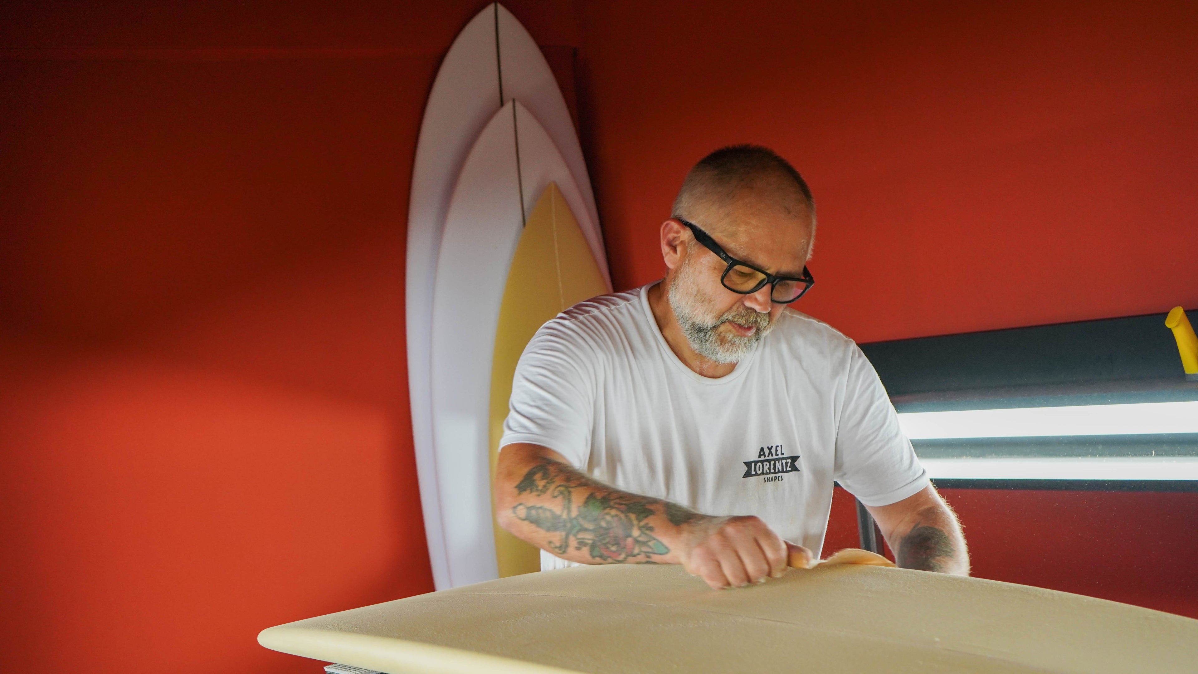 Man working on a surfboard in a workshop with a red wall and another surfboard in the background.